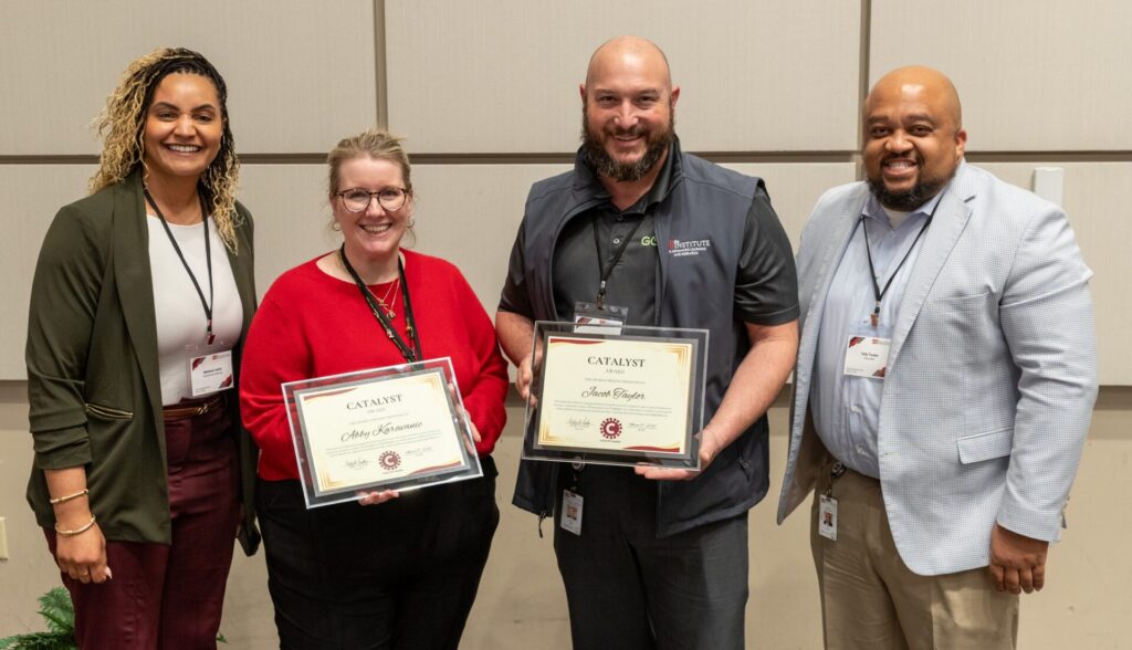 A group of four people smiles at the camera. The two in the middle are holding awards.