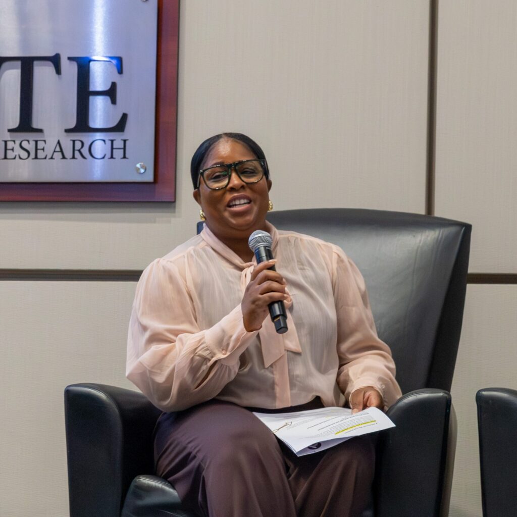 A woman speaks into a microphone while sitting in a chair on a stage. 